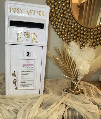 A decorative white post box with royal insignia, next to a mirror and dried plants.